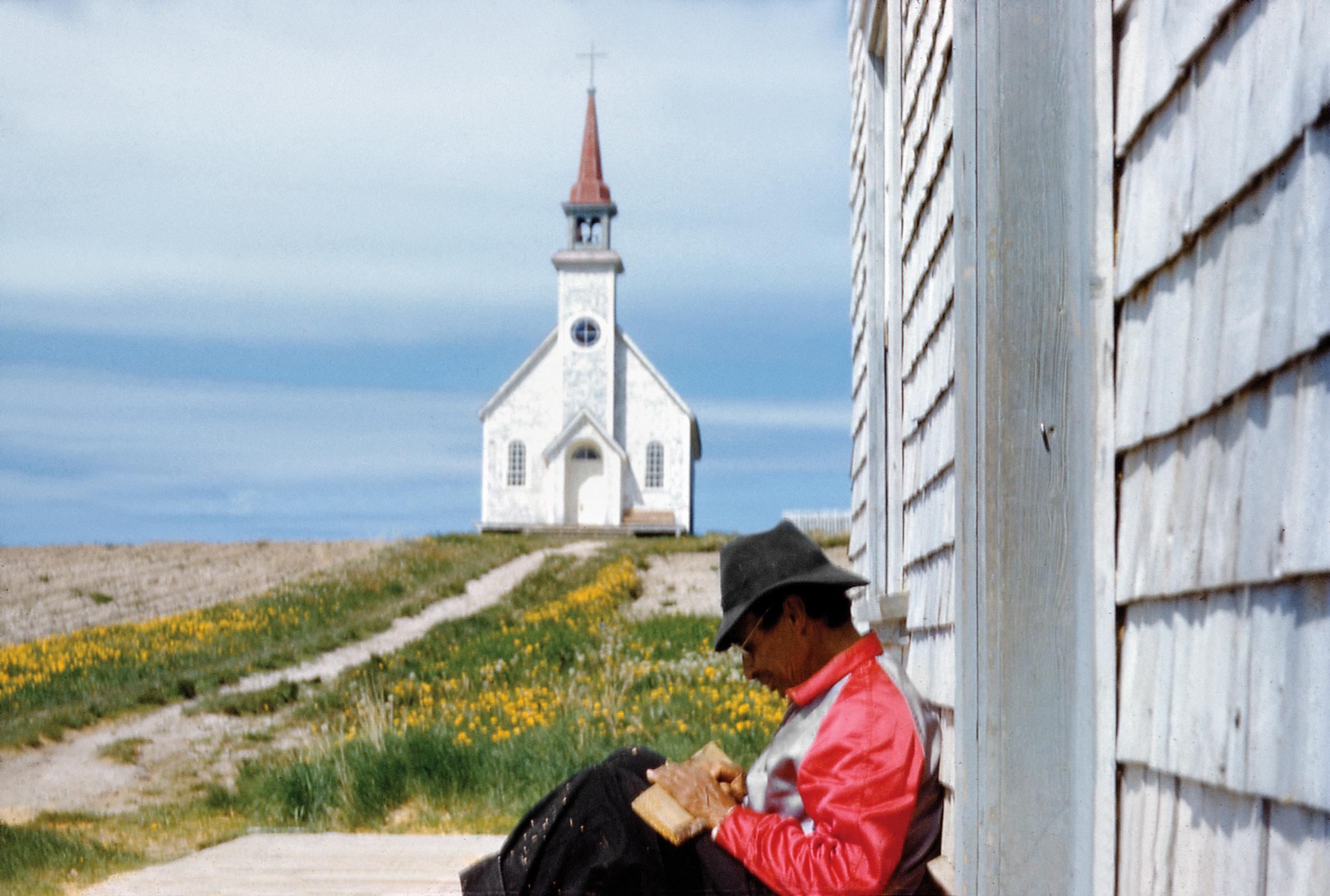 John Macfie, Man sitting against a building with a church in the background, Sandy Lake, c.1952. Archives of Ontario, John Macfie fonds, C330-14-0-0-103